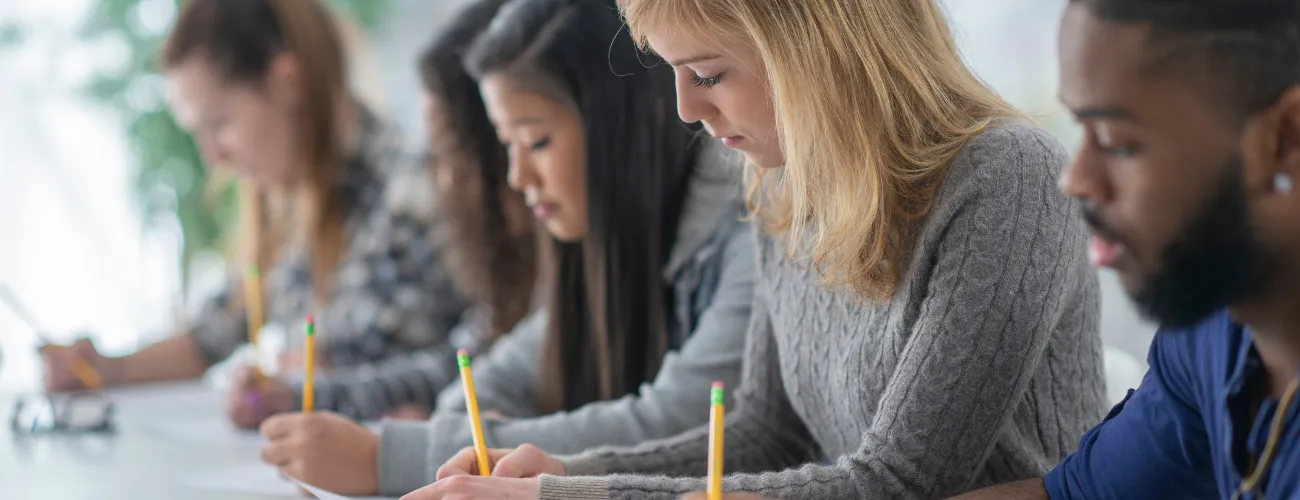 Five students sitting at a desk taking a test with pencils