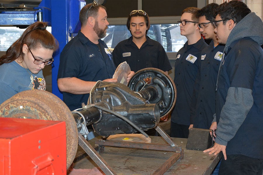 Automotive career and technical education students listening to a teacher while standing in front of parts of a car