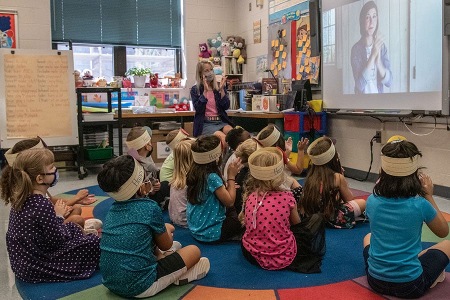 A group of young students sit on the floor in front of their teacher while watching a video