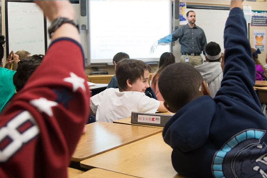 Middle school students sitting at their desks in a classroom with some students in the foreground raising their hands to be called on