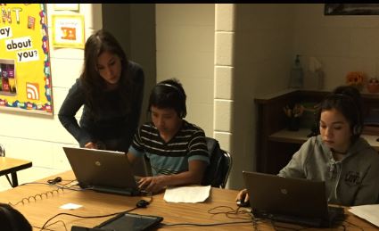 Two students sitting at the same table in front of computers with a teacher helping one of the students