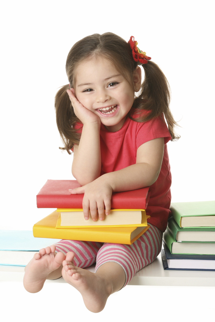 A smiling toddler girl sitting with her hand under her chin. She is holding 3 books in her lap. There are 4 more books by her side.