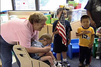 A classroom showing two boys with hands on their chest facing the flag of USA. A teacher helping another boy in a chair to raise the US flag