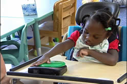 A student sitting in an adapted chair pressing the button on a big MACK switch.