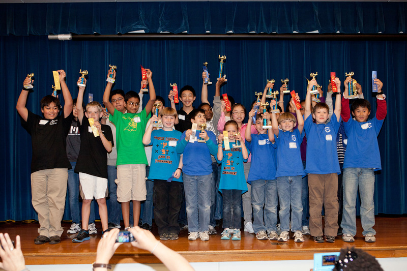 A large group of students standing on a stage. Each child is holding a trophy high above their head.
