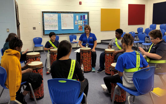 educator and several students seated in a circle beating on a drum