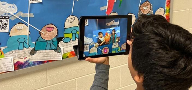 Male student holding a tablet up to a bulletin board. 