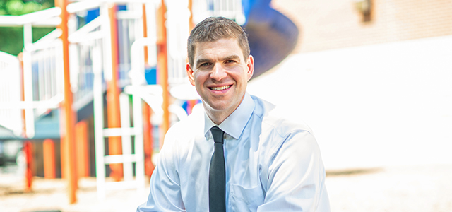 waist-up photo of seated school principal Dr. Andy Jacks in shirt and tie with a school playground in the background