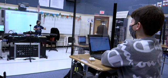 Student at desk behind plexi-glass with teacher in classroom