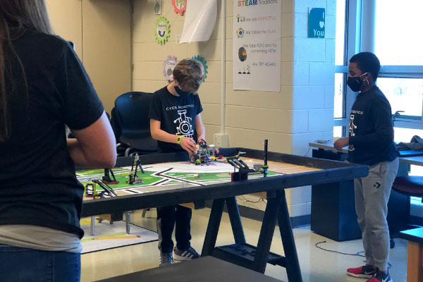 Several students wearing face coverings working on robotic parts at a table in a classroom