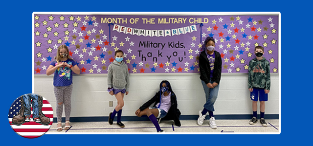 Five students with face coverings standing and sitting in front of a patriotically decorated bulletin board in a school hallway