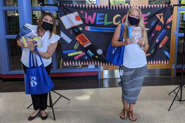 full length photo of the River Oaks ES principal, reading teacher, and assistant principal with face coverings standing safely distanced holding books at the school in front of a welcome banner
