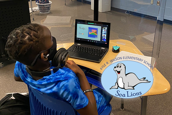 Student sitting at desk in front of laptop with headphone. Kyle Wilson sea lion logo in bottom right corner.