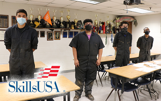 Four automotive students in masks standing in a classroom with a shelf of trophies and plaques behind them