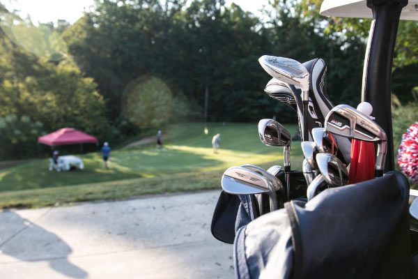Foreground view of the back of a golf cart with the top of a golf bag and clubs sticking out, with the green and a hole in the background.