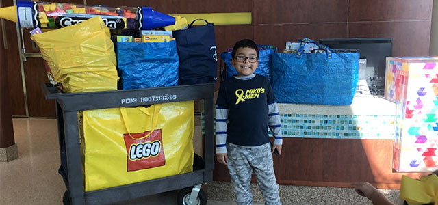 Full length photo of Niko, a Haymarket Elementary School student, smiling and standing in front of a cart piled high with LEGOs donated for the school's LEGO Drive