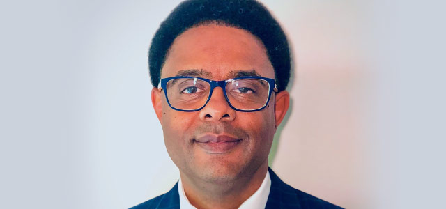 Smiling head-and-shoulder photo of Corey Harris, who has short black hair and black-framed glasses and is wearing a dark suit collar, white shirt, and bow tie, image background has a white halo effect around his head that fades slightly grey at the edges