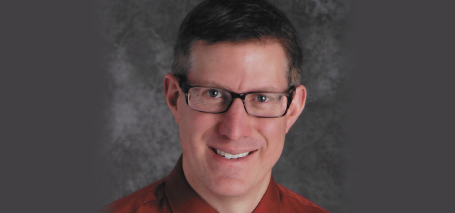 Smiling headshot image of Reed Gillespie with short brown hair and glasses and a red shirt collar