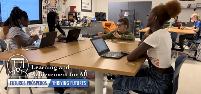 Sudents pictured with laptops sitting around a table in classroom.