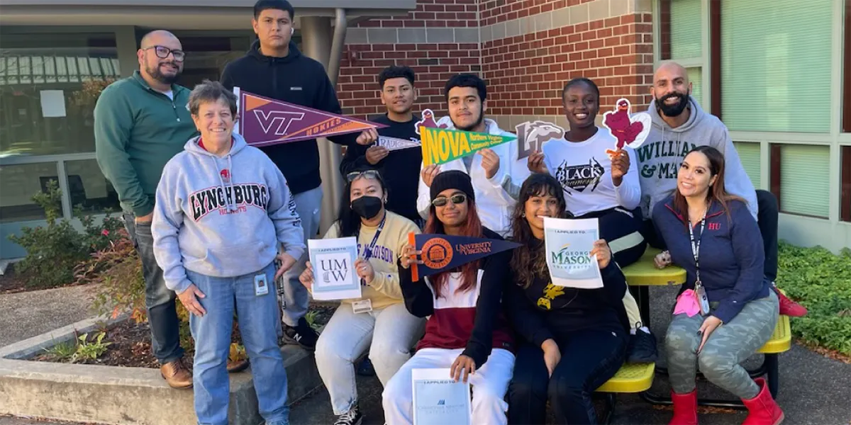 Diverse group of Freedom High School students and staff sitting outside the school holding signs for colleges
