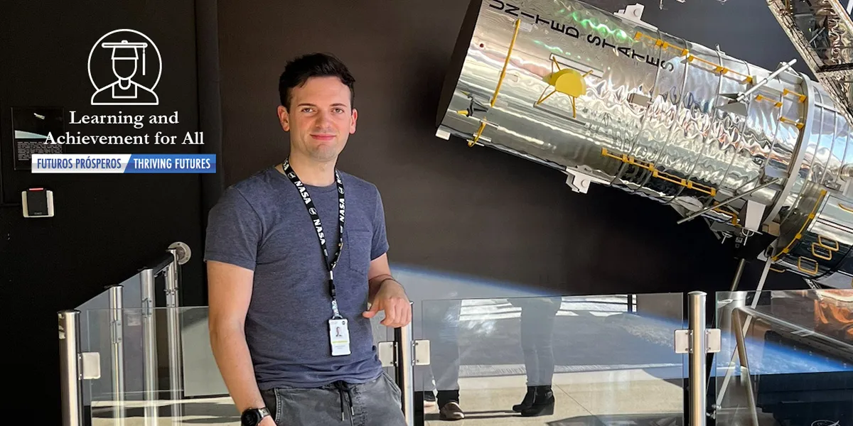 waist-up photo of 2012 PWCS graduate Johnathan Brendle in a soft blue t-shirt leaning against a glass display case  in front of a Hubble Space Telescope scale model