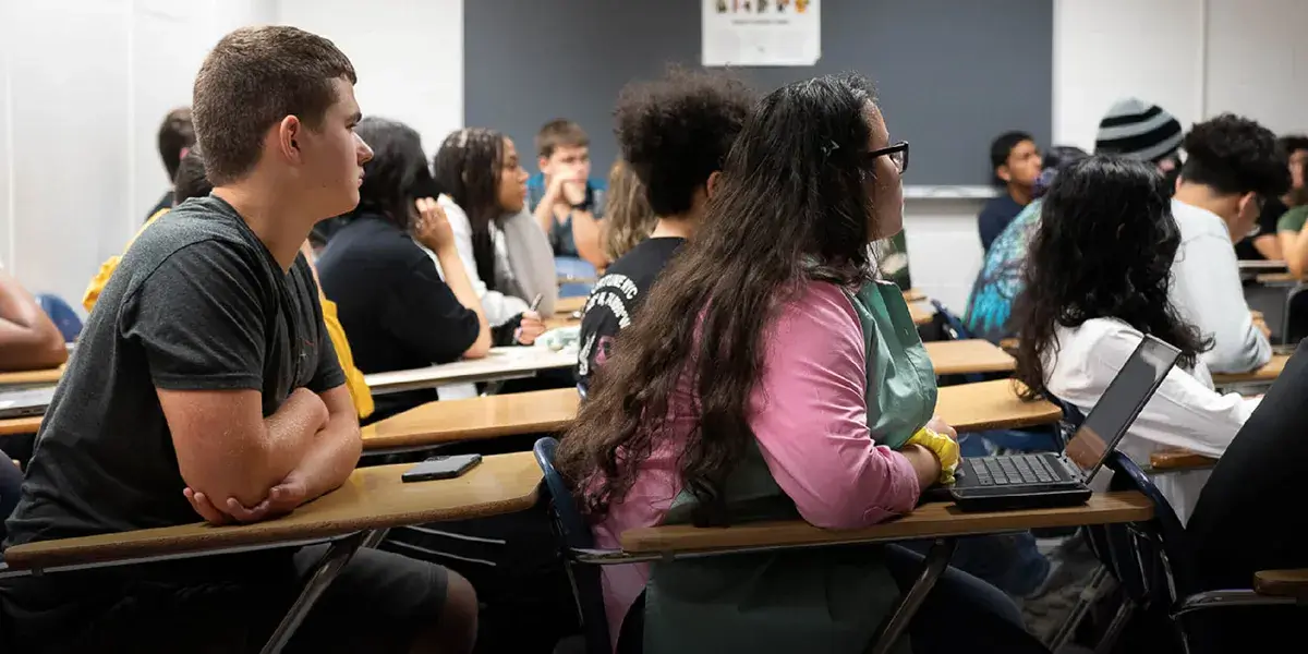 Side view of a classroom of high school students sitting at their desks with their attention focused toward the front of the room