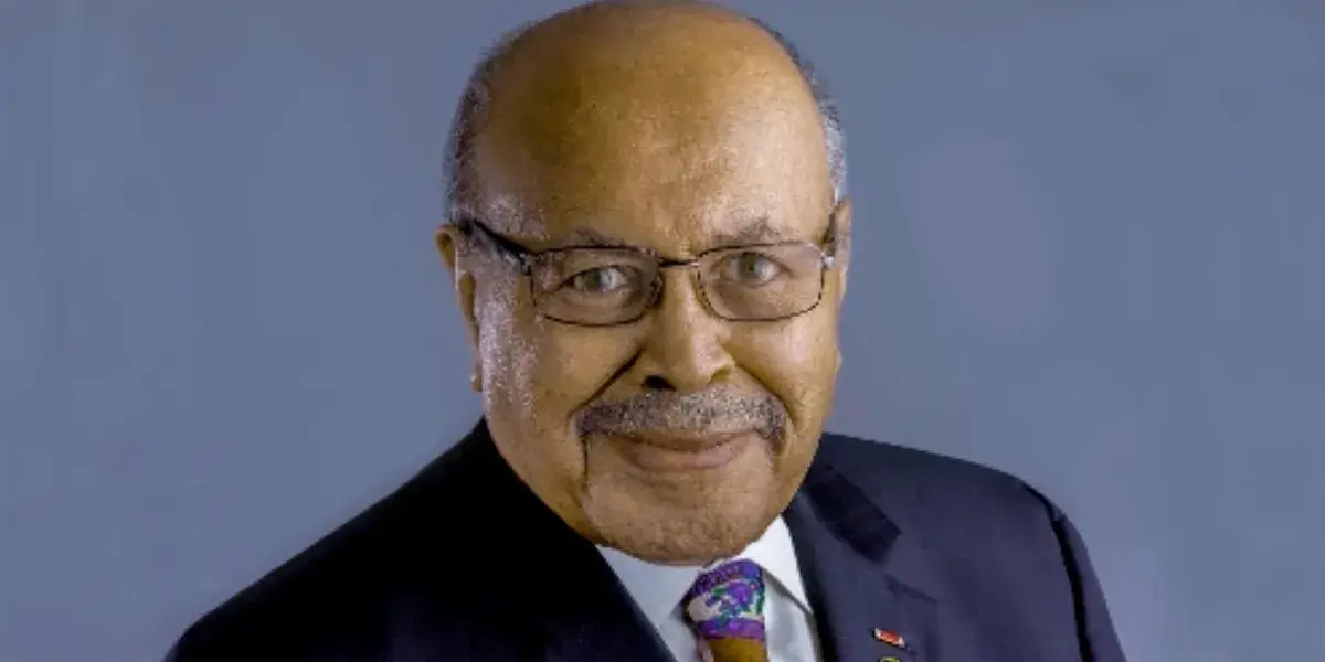 Smiling headshot photo of Dr. George M. Hampton, head and shoulders, balding but with a ring of grey hair and a mustache, wearing a navy suit and colorful, purple tie, with pins on the lapel.