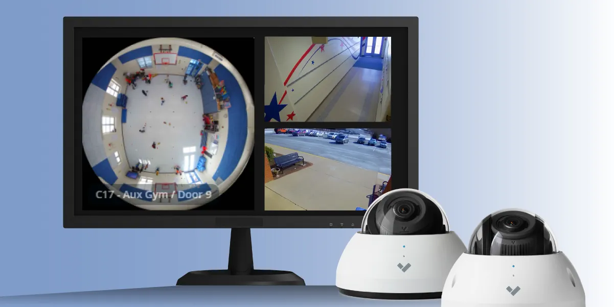 Picture of a computer monitor standing on a surface in front of a light blue background and displaying three different viewpoints of school areas as seen from a camera receiver. In the foreground of the picture are two base Verkada closed-circuit television (CCTV) system cameras sitting on the surface in front of the monitor.