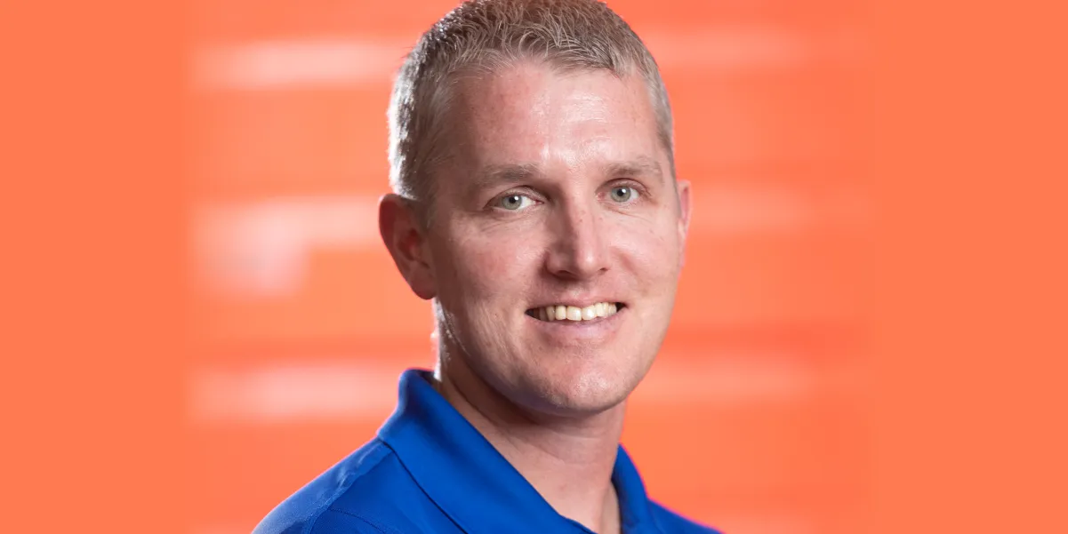 Smiling headshot photo of Michael Payne, director of student activities at Gar-Field High School, showing the collar of a royal blue polo shirt and on an orange background