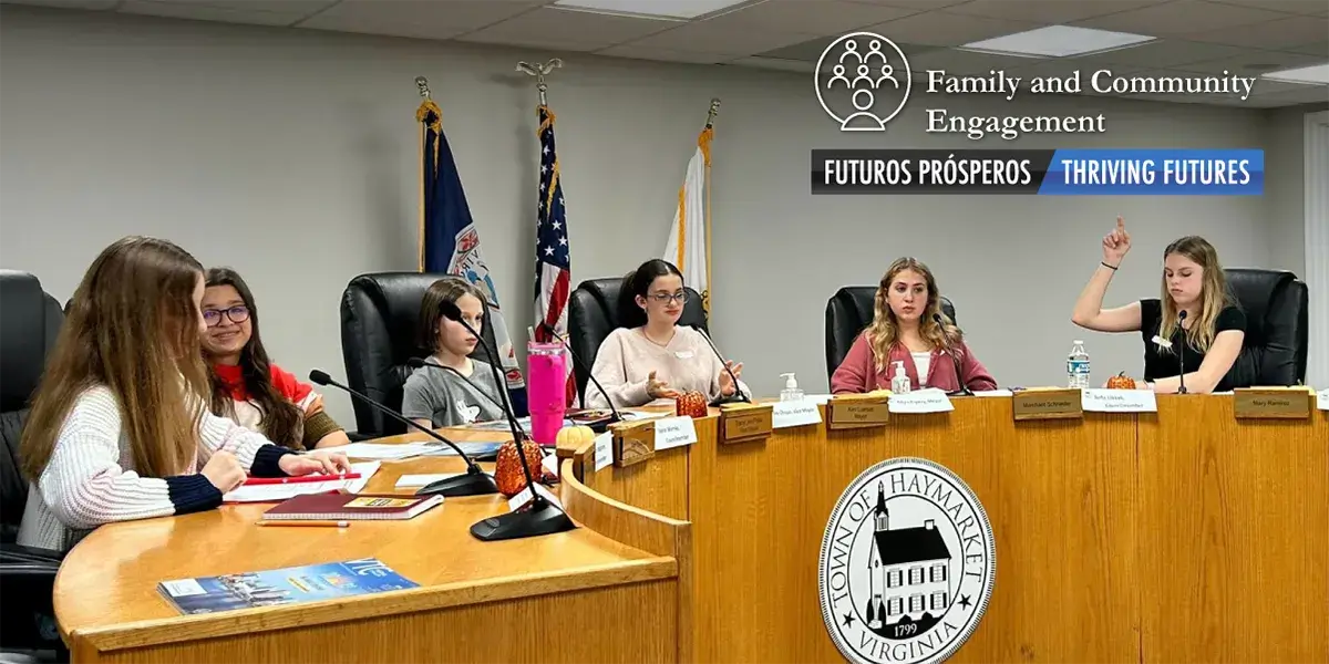 Six middle school students sitting on the dais that displays the town of Hayrmarket emblem on the front. The students are conversing with each other as they are holding a meeting, with three flags in the background.