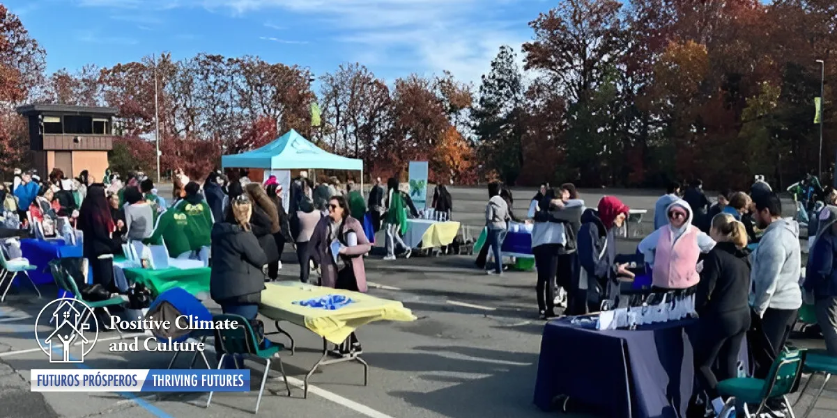 Photograph outdoors on a beautiful sunny day with blue sky and clouds and trees in the background and a fair event being held in a school parking lot with people, tents, and activities in the foreground