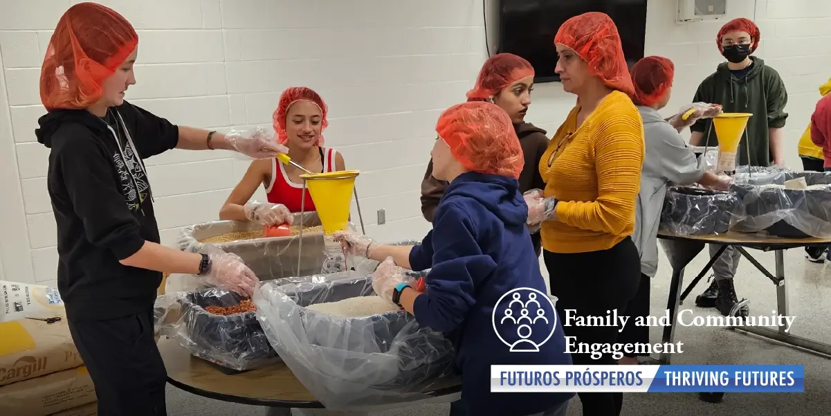 Photograph of about eight people who are wearing hair nets and appear to be part of a bigger group of teens and adults who are standing and sitting at school tables arranging food packages. Text in the bottom right of the image reads "Family and Community Engagement. 