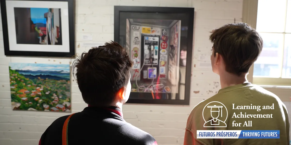 Photo image that shows two dark haired teen boys’ heads and shoulders from behind as they gaze at a framed piece of artwork on a painted brick wall – there is light shining on them coming from a window to the right of the art and some of their facial features are visible in the reflection of the glass in the framed artwork.  