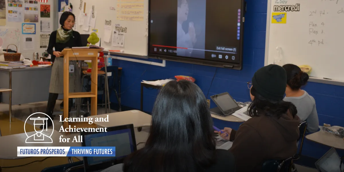 Photo of a classroom, showing desks in the foreground and from behind, the backs of three teen students heads. The students are facing a dark-haired adult female teacher in a skirt and long-sleeve black turtleneck with a scarf around her neck and standing a podium looking down at a textbook. On the wall to the teachers left is a dry erase board and in from of that is a e-whiteboard with a video showing a Korean teen singing and holding a microphone. Text and image overlay on the bottom left corner of the graphic shows the outlined figure of a student wearing a graduation cap and the text “Learning and Achievement for All, Futuro Prosperos/Thriving Futures”