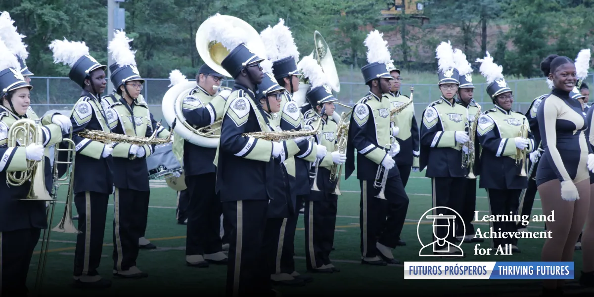 A photo of about 30 high school students in colorful marching band uniforms and holding large band instruments outdoors on a background of grass and trees.