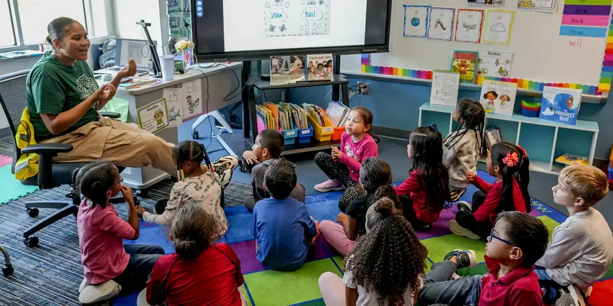 Elementary school teacher sitting in a classroom while talking to a diverse group of students sitting on the floor