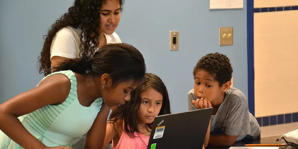 Four students (three standing, one sitting) in front of a laptop in a school classroom