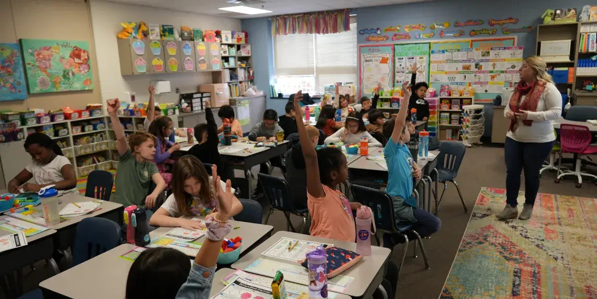 Elementary school teacher standing in front of a classroom of students seated at their desks. Many of the students are holding up their hand waiting to be called on.