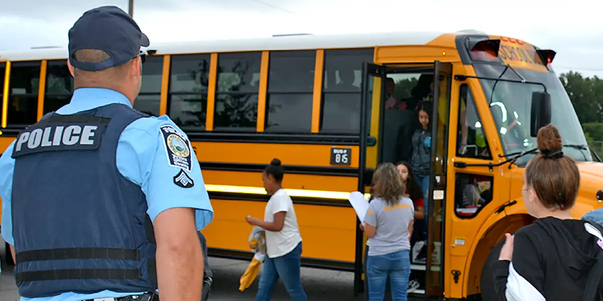 School resource officer standing in front of a school bus while students arrive at school