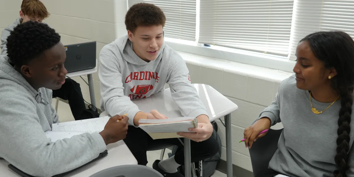 Three Gainesville High School students sitting at desks in a classroom actively engaged with each other