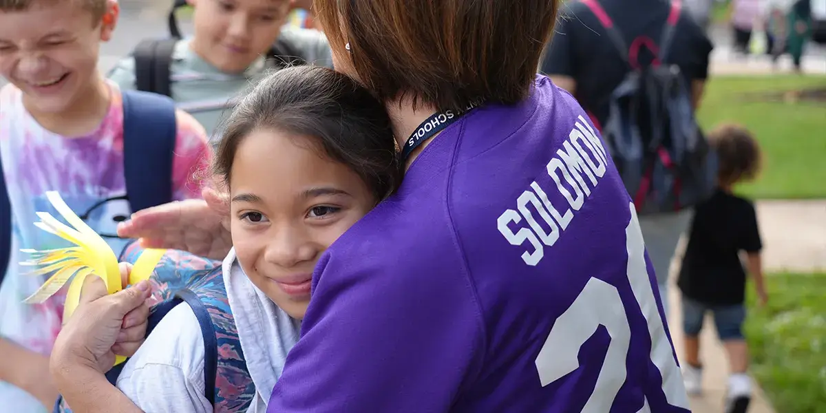 A Cedar Point Elementary School teacher and student hug while students enter the building on the first day of school