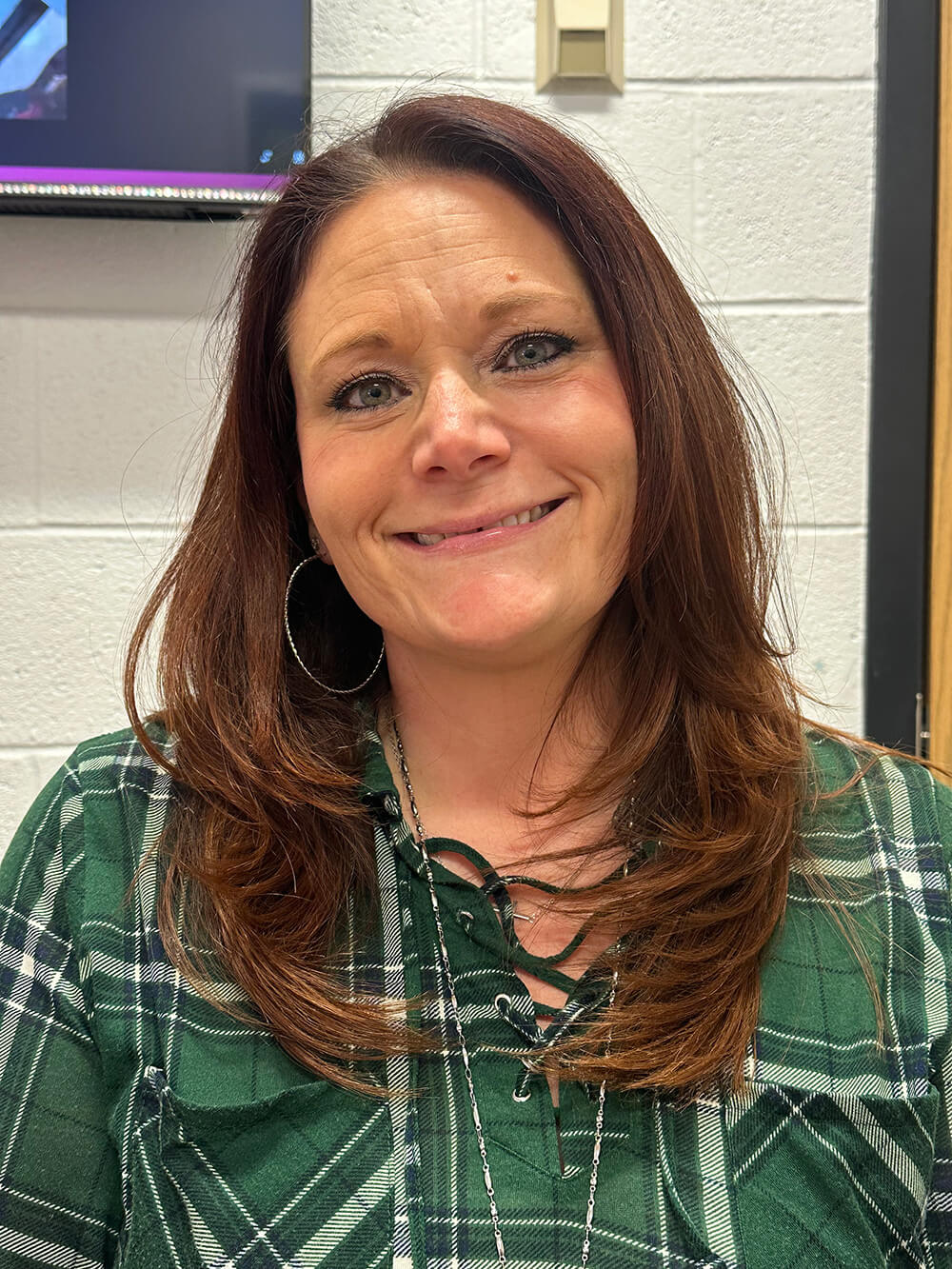 A smiling woman with long brown hair wearing a green plaid shirt and large earrings.