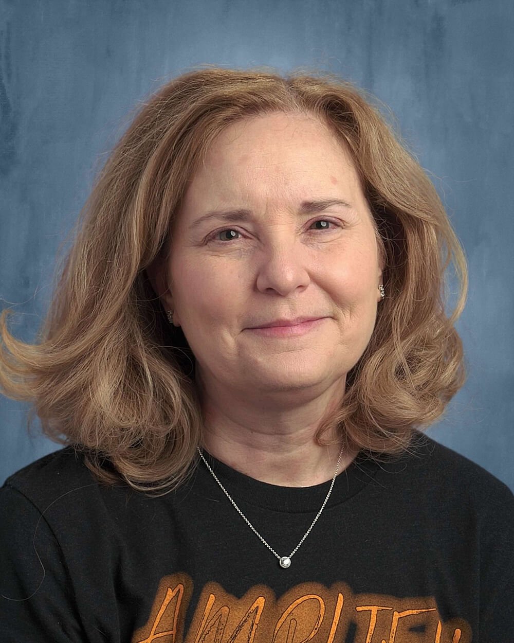 A smiling woman with shoulder-length wavy hair wearing a dark t-shirt and a necklace, set against a blue background.
