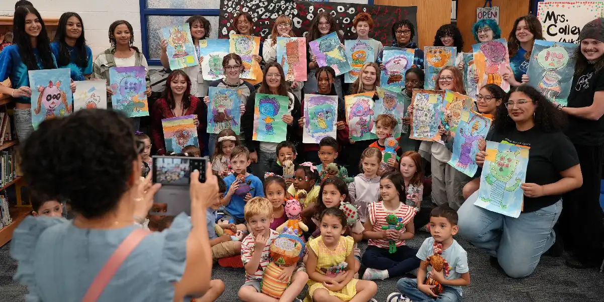 A group of elementary and high school studentsholding up their colorful artwork, celebrating creativity together in a bright classroom environment.