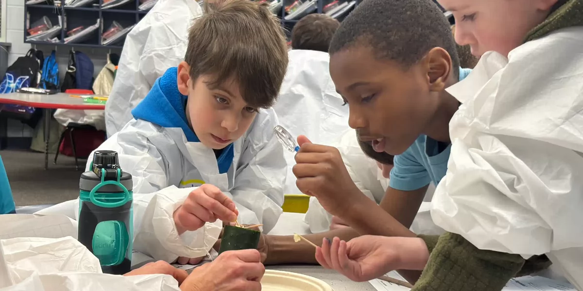 Three students surrounding a science experiment and one of them is holding a magnifying glass.