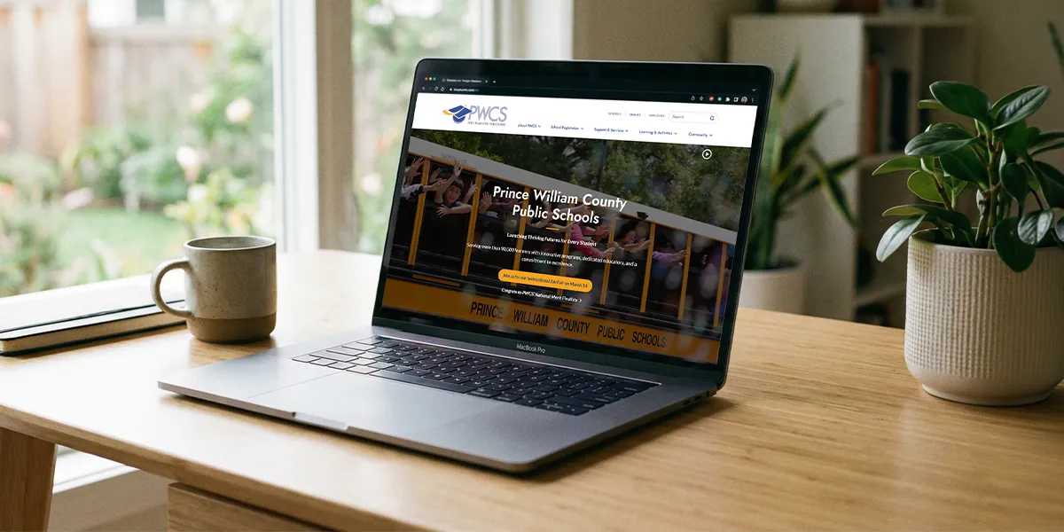 Laptop on a desk displaying the Prince William County Public Schools website next to a coffee mug and a potted plant.