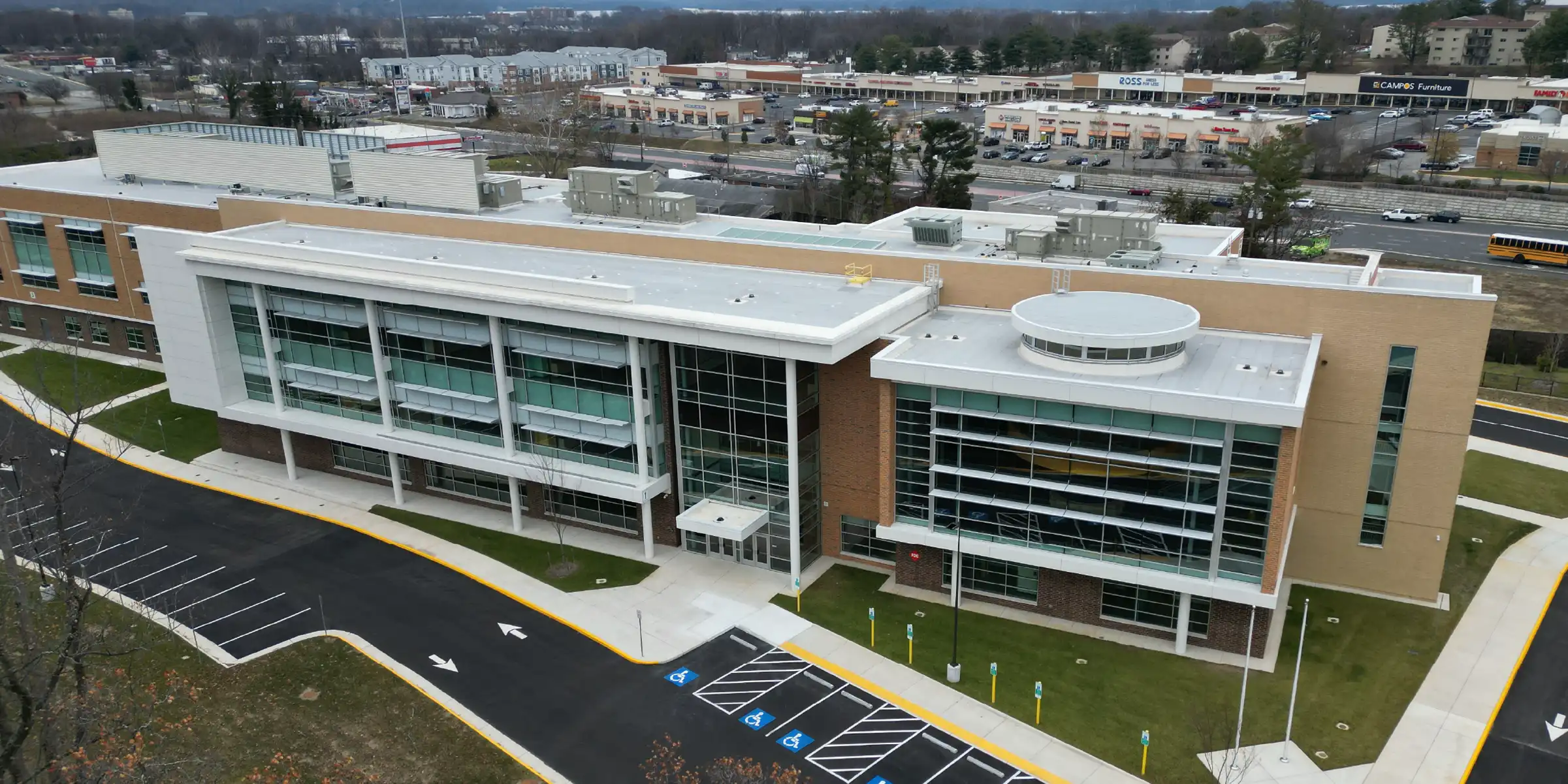 Aerial view of a modern multi-story campus building with a glass-front entrance and a curved rooftop feature beside an empty parking lot.
