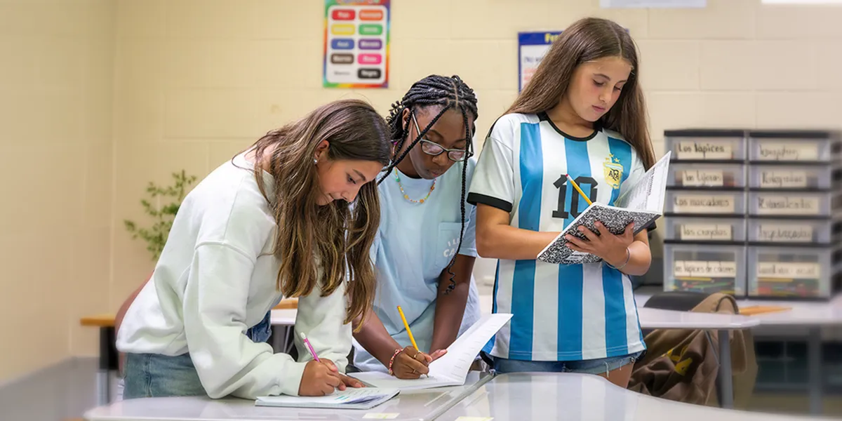 Three students work together in a classroom, writing on papers at a table while another reads from a notebook.