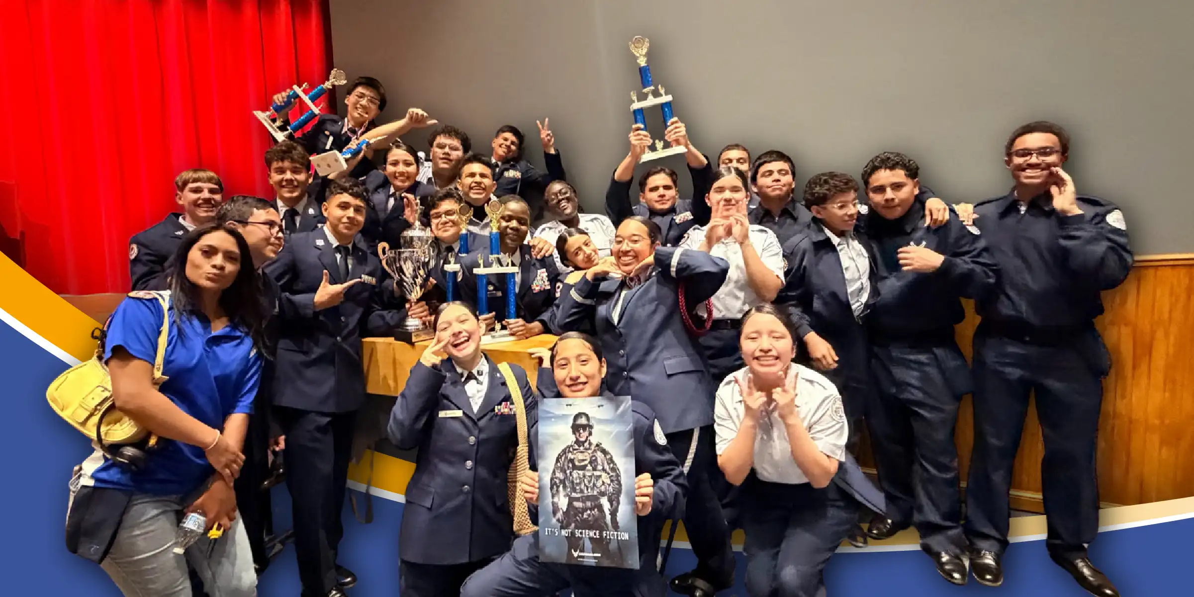 Group of students in uniform pose on a stage holding trophies and cheering in celebration.