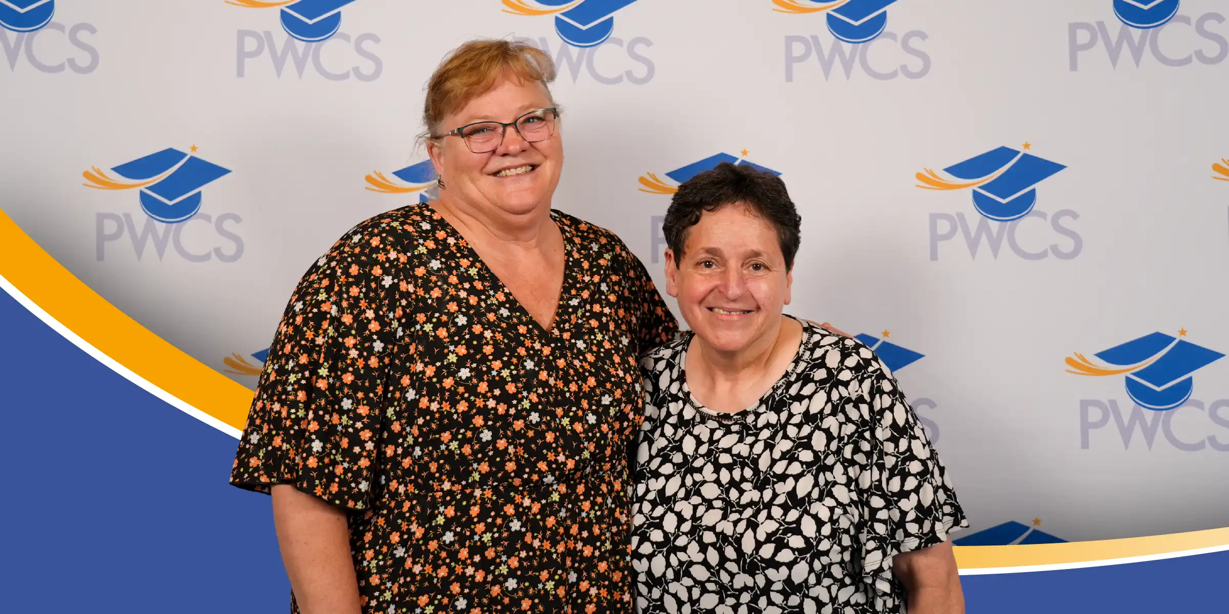 Two women smiling and posing in front of a PWCS step-and-repeat backdrop with graduation cap logos.